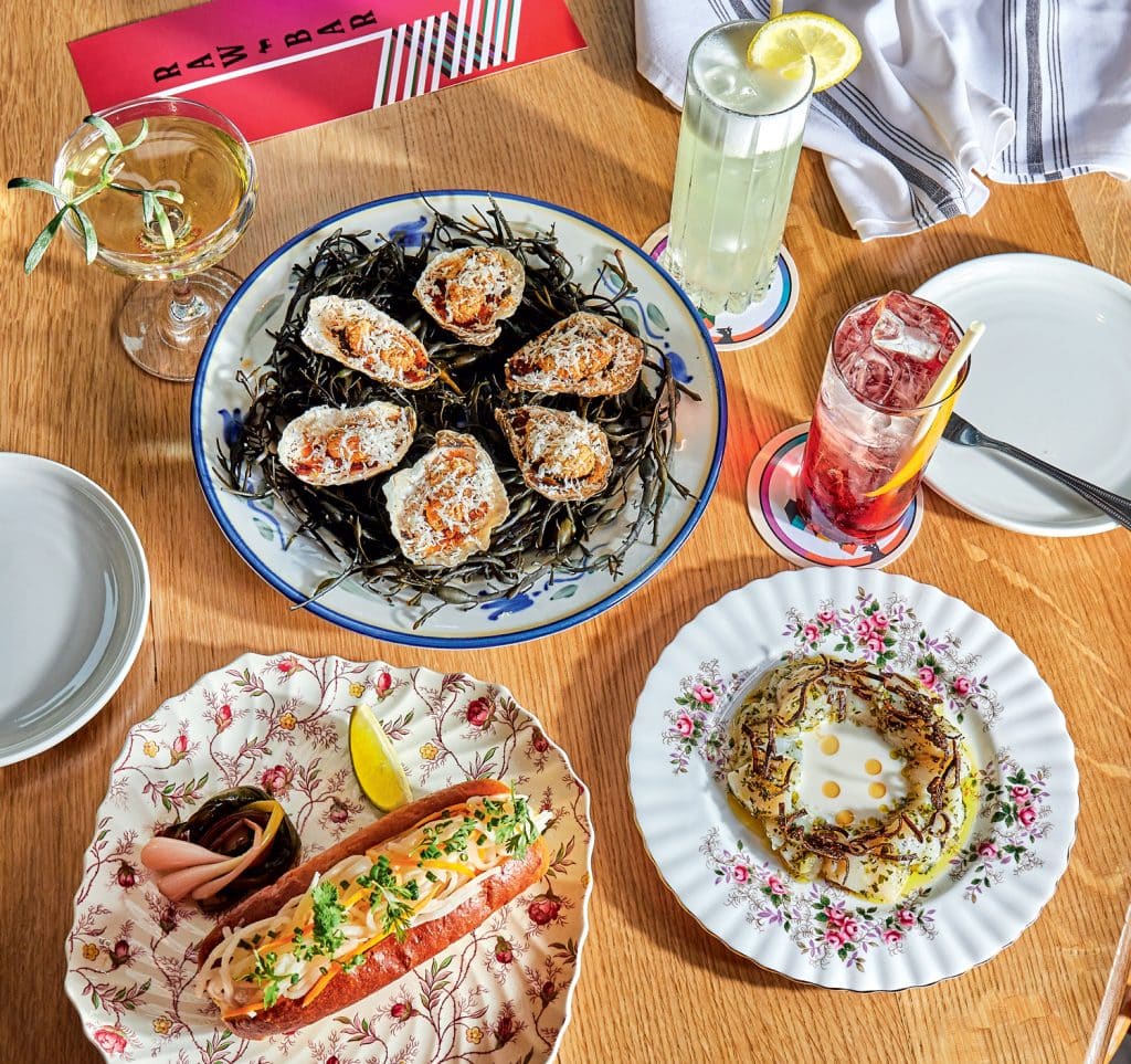 A table with oysters on seaweed, a lobster roll, a potato dish, and two cocktails on floral and plain plates.