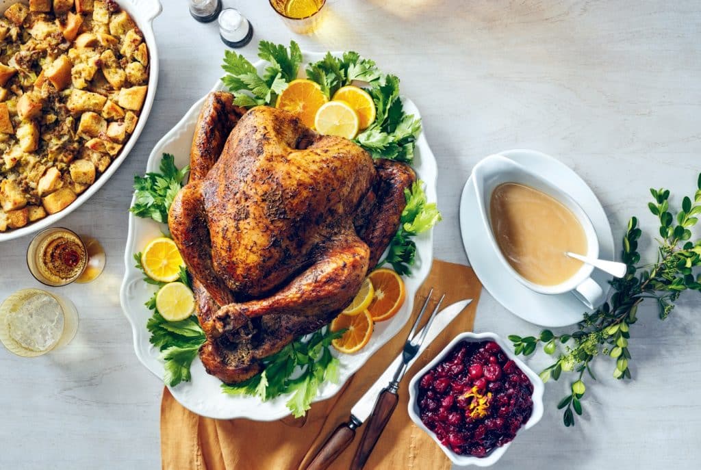 Roast turkey garnished with lemon slices and herbs, surrounded by stuffing, cranberry sauce, and gravy on a table.