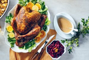 Roasted turkey on a platter with orange slices and herbs, surrounded by bowls of cranberry sauce and gravy, with serving utensils and a sprig of greenery.