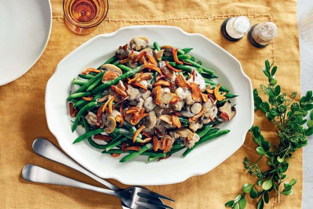 A white platter of green beans topped with sautéed mushrooms and crispy onions on a yellow tablecloth, with cutlery and a small green plant beside it.