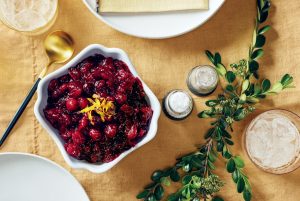 A bowl of cranberry sauce garnished with orange zest is on a table with a gold spoon, salt and pepper shakers, a green leaf branch, and a glass with ice on a mustard-colored tablecloth.