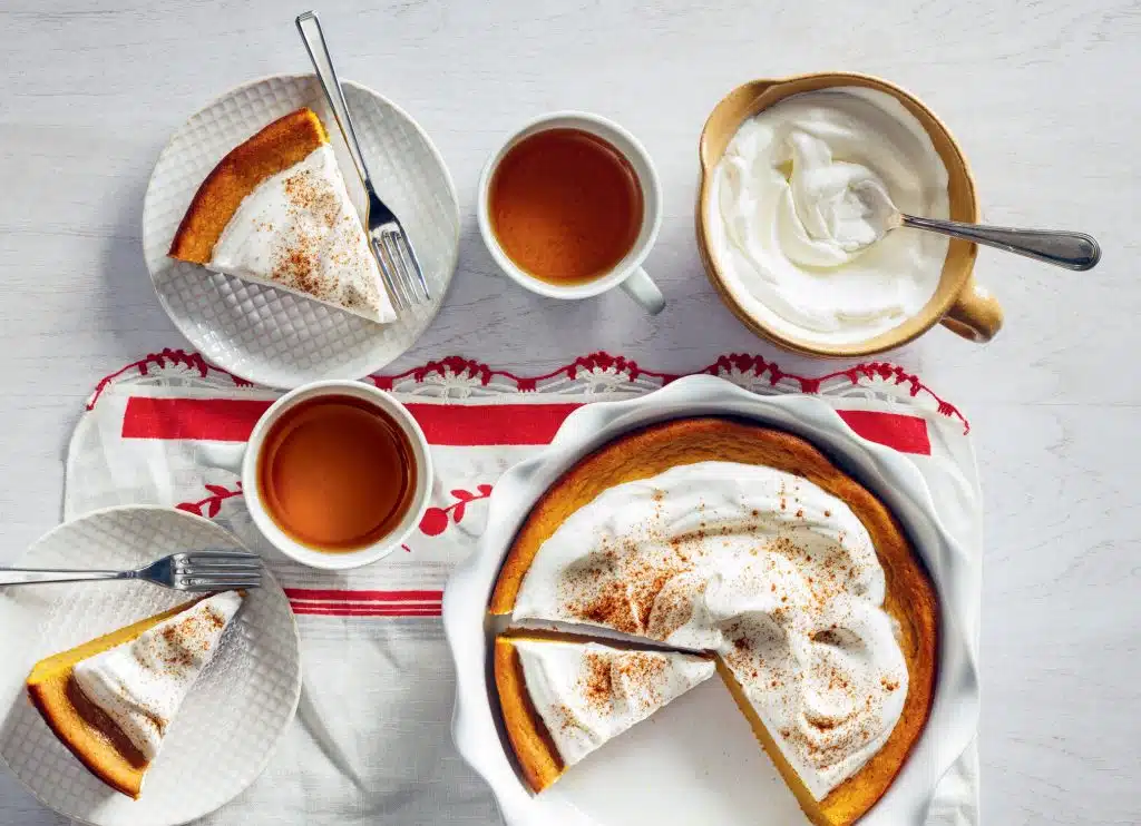 A table with pumpkin pie slices, two cups of tea, a bowl of whipped cream, and a whole pie on a red-and-white cloth.
