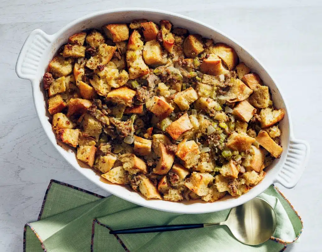 A white oval dish filled with golden-brown stuffing sits on a table with green napkins and a gold spoon.