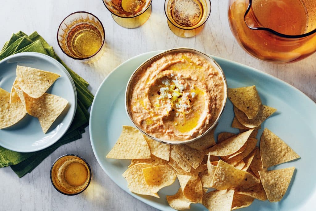 A bowl of feta dip with olive oil garnish on a plate with tortilla chips. Accompanied by a stack of napkins, an empty plate, several glasses, and a pitcher on a white surface.