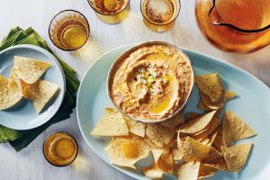 A bowl of feta dip with olive oil garnish on a plate with tortilla chips. Accompanied by a stack of napkins, an empty plate, several glasses, and a pitcher on a white surface.