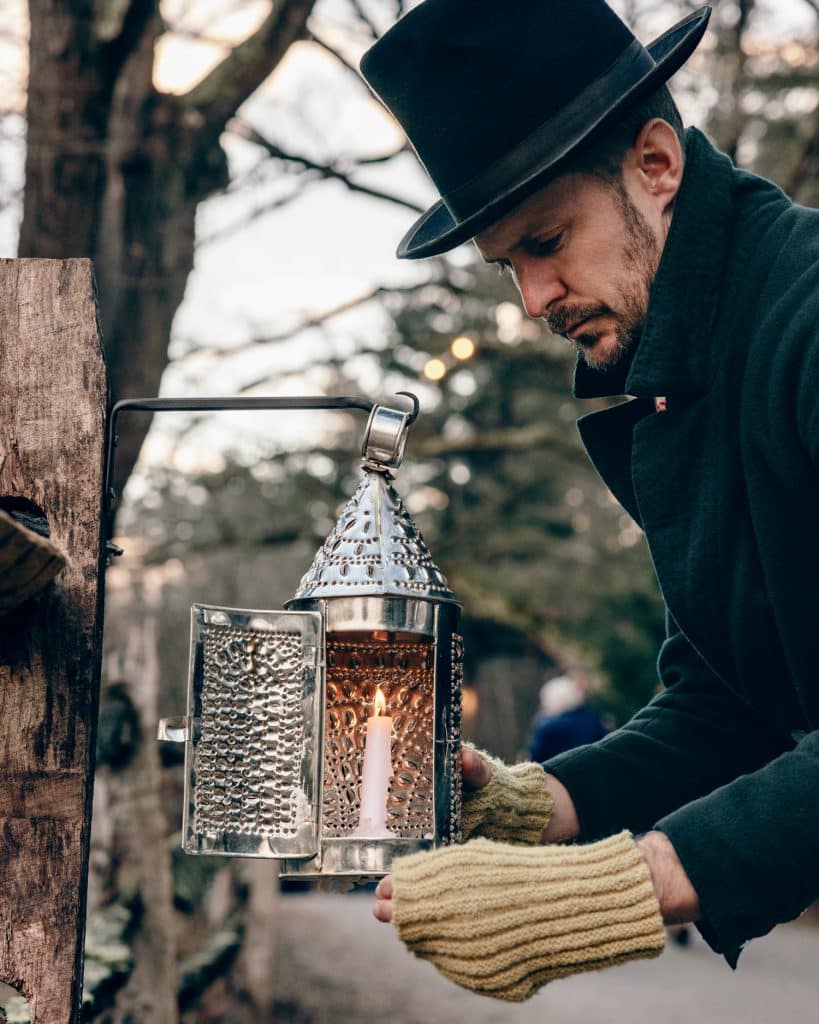 A person in a black hat and coat with green mittens adjusts a lit candle inside a decorative lantern outdoors.