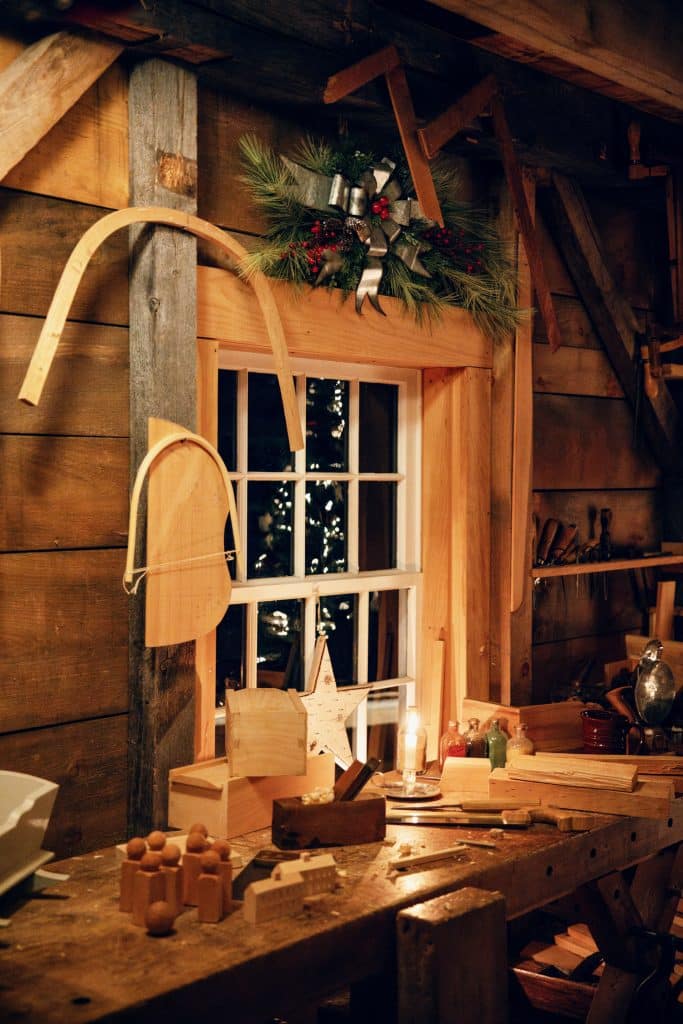 A cozy woodworking shop with a wooden workbench, scattered tools, and small wooden crafts. A window is adorned with a seasonal wreath and a candle, providing soft illumination.