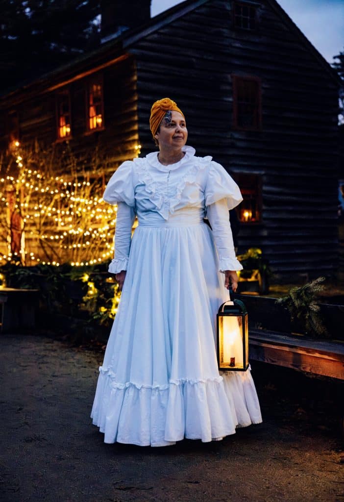 Person in a white historical dress with a yellow head wrap holds a lit lantern, standing outside a wooden building adorned with string lights at dusk.