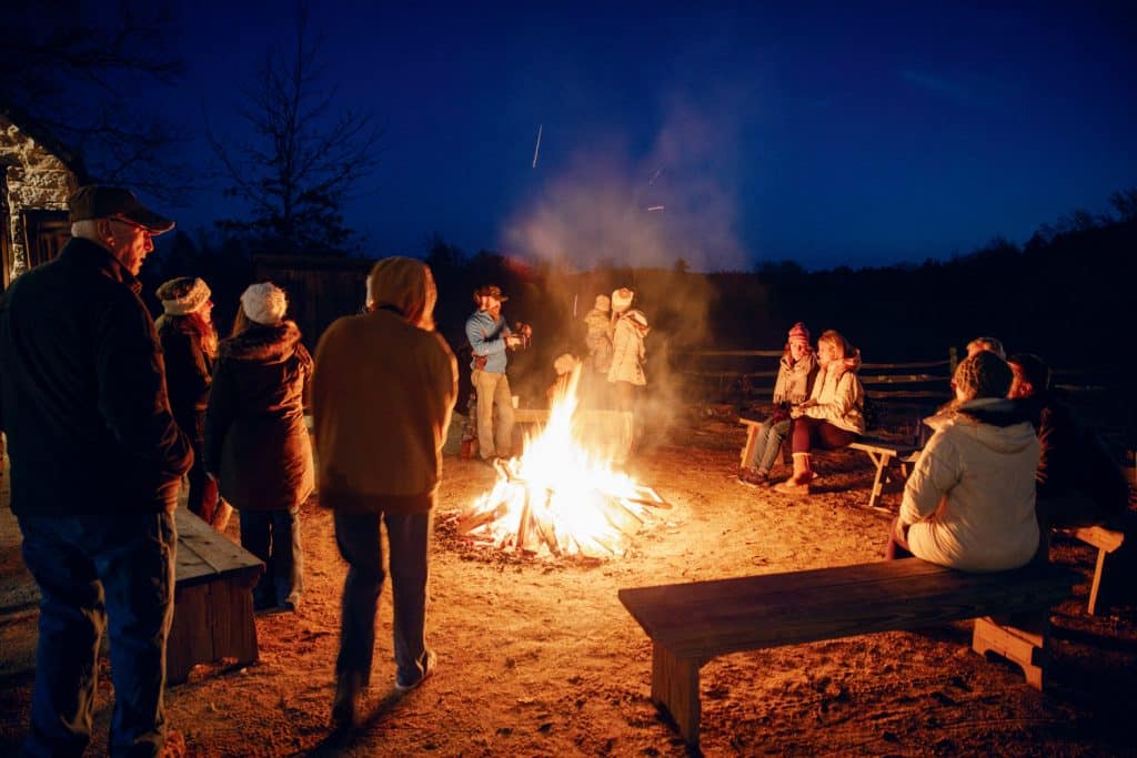 People gathered around a campfire at night, wearing warm clothing and sitting on wooden benches. The sky is dark with visible stars.