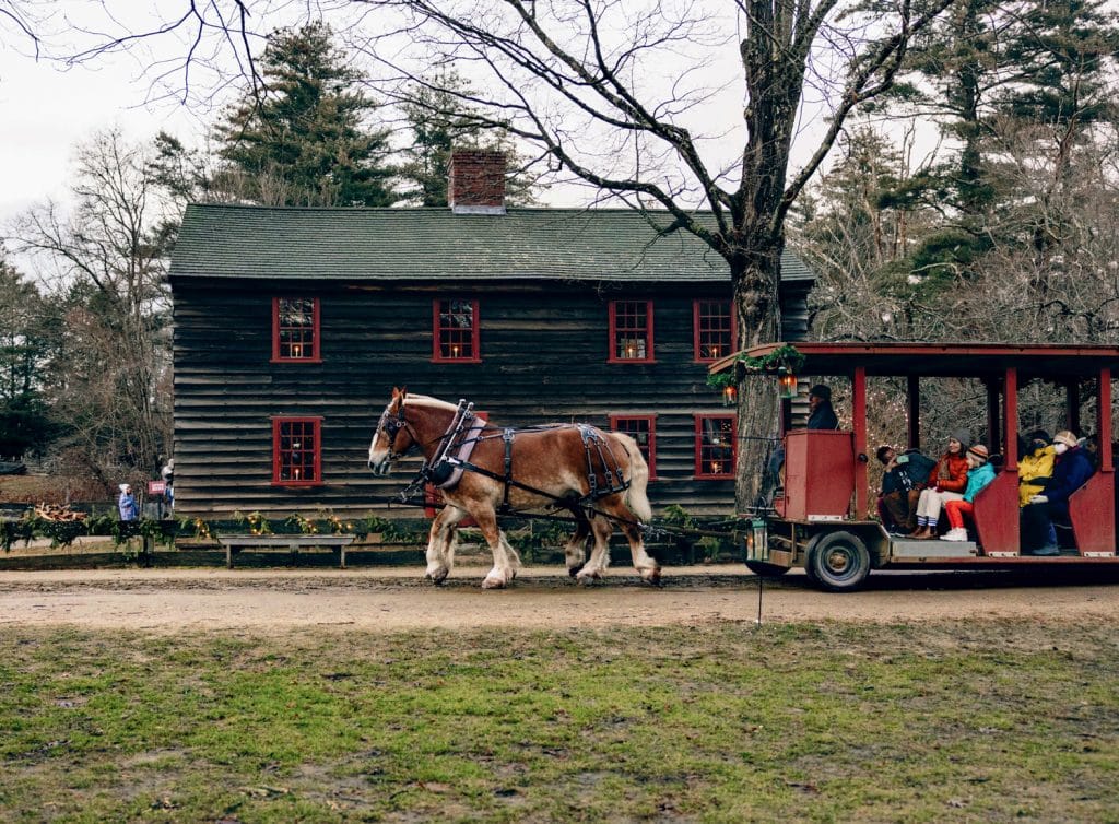 Horse-drawn carriage with passengers rides past an old wooden house on a cloudy day.