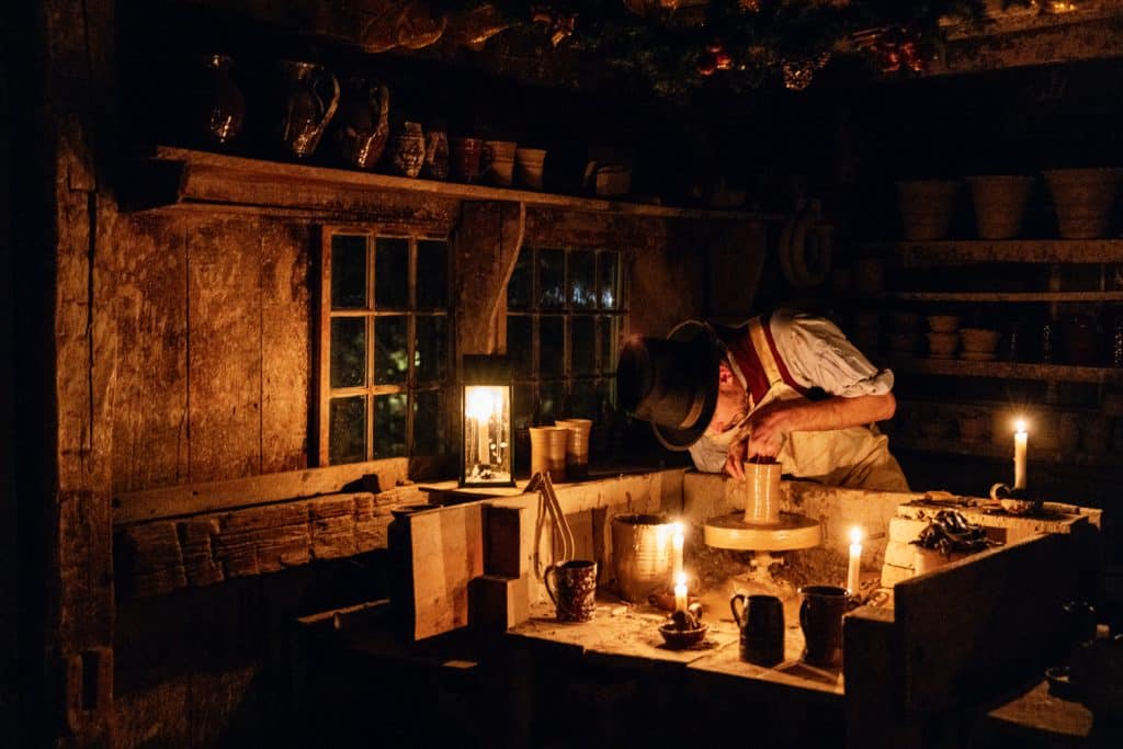 A person in old-fashioned attire shapes clay on a potter's wheel, surrounded by candles and pottery in a dimly lit workshop.