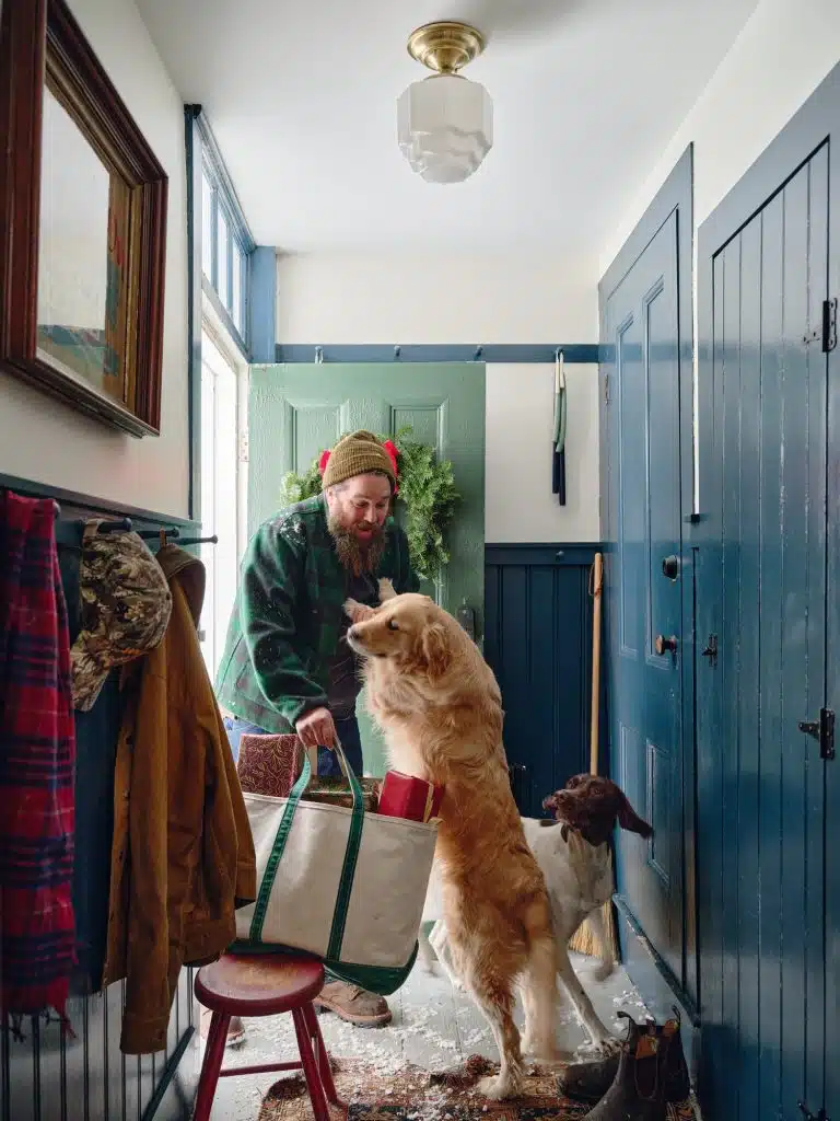 A man wearing a beanie holds a tote bag in a blue-paneled foyer. A golden retriever jumps up as another dog stands nearby. Coats hang on the wall and snow is scattered on the floor.