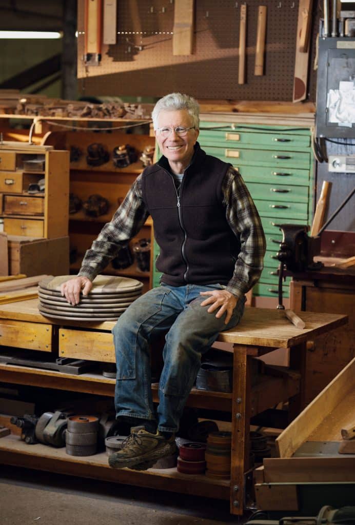 Older man sitting on a table in a woodworking shop, wearing glasses, a plaid shirt, and a black vest. Background includes shelves with tools, wooden boards, and a green drawer cabinet.