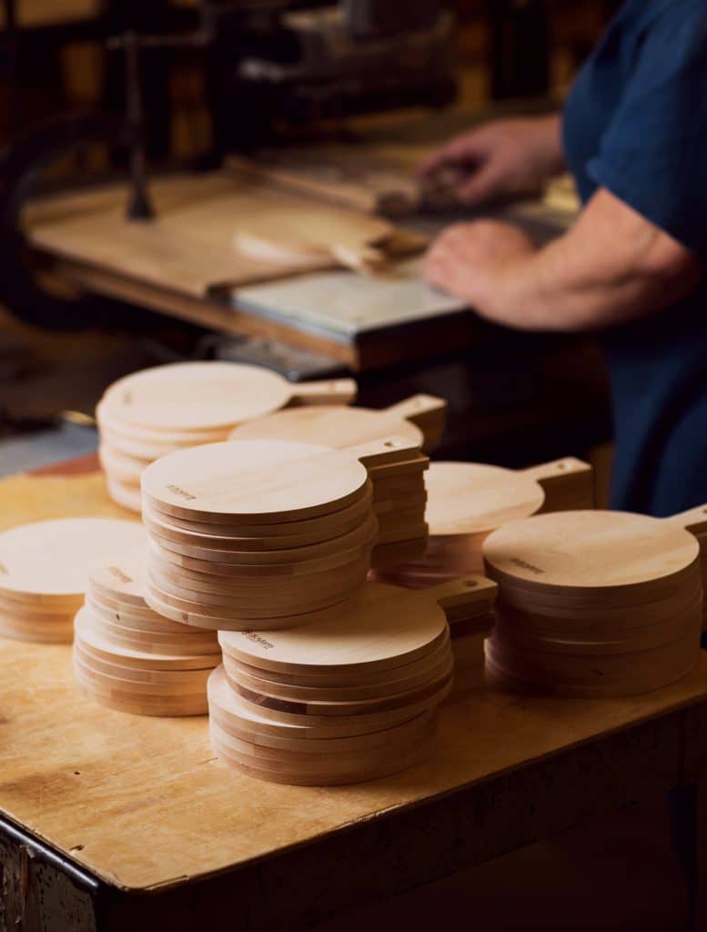 Stacks of unfinished wooden cutting boards on a table in a workshop, with a person working in the background.