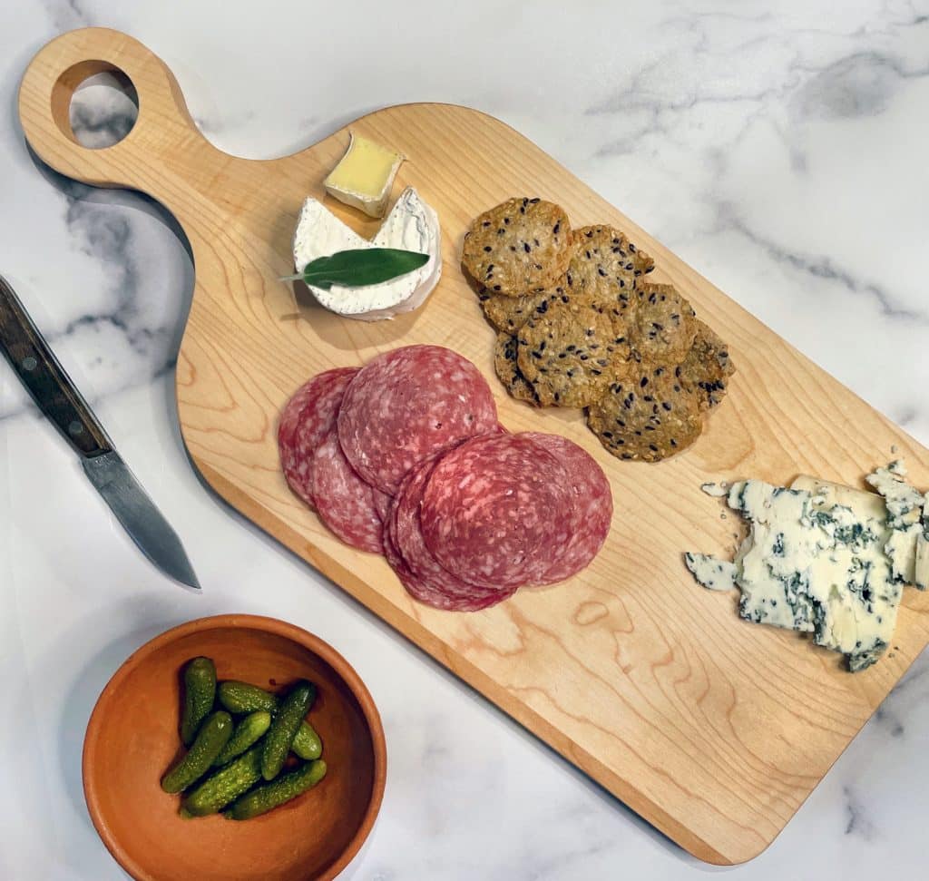 A wooden board with sliced salami, blue cheese, brie with leaf, crackers, and a small bowl of pickles. A knife is placed beside the board.