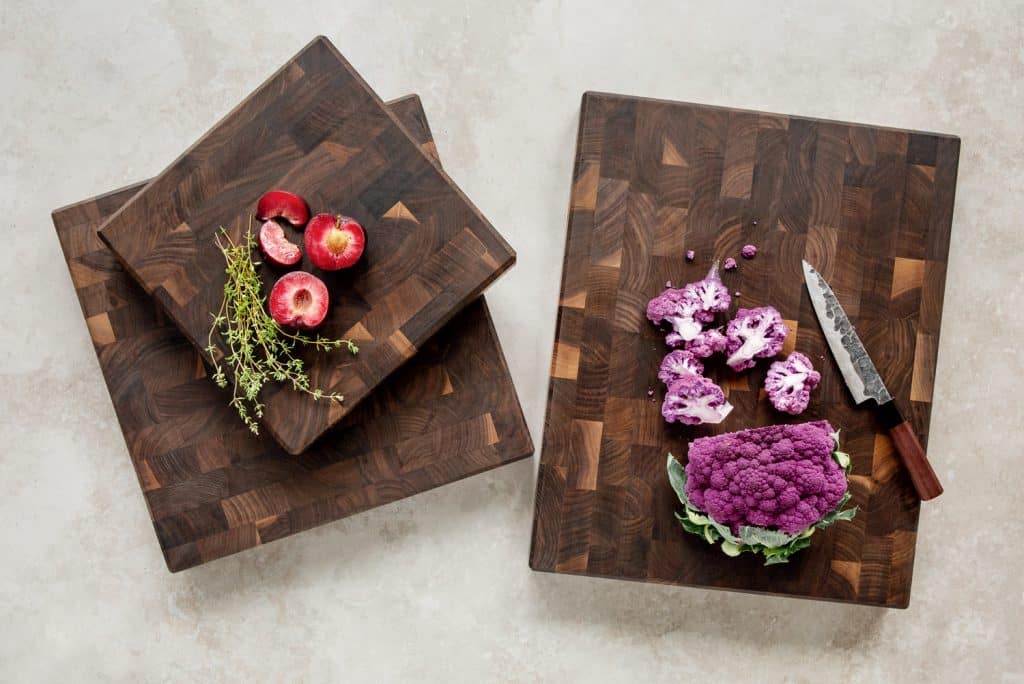 Cutting boards with plums, thyme, and a knife beside chopped and whole purple cauliflower on a light surface.