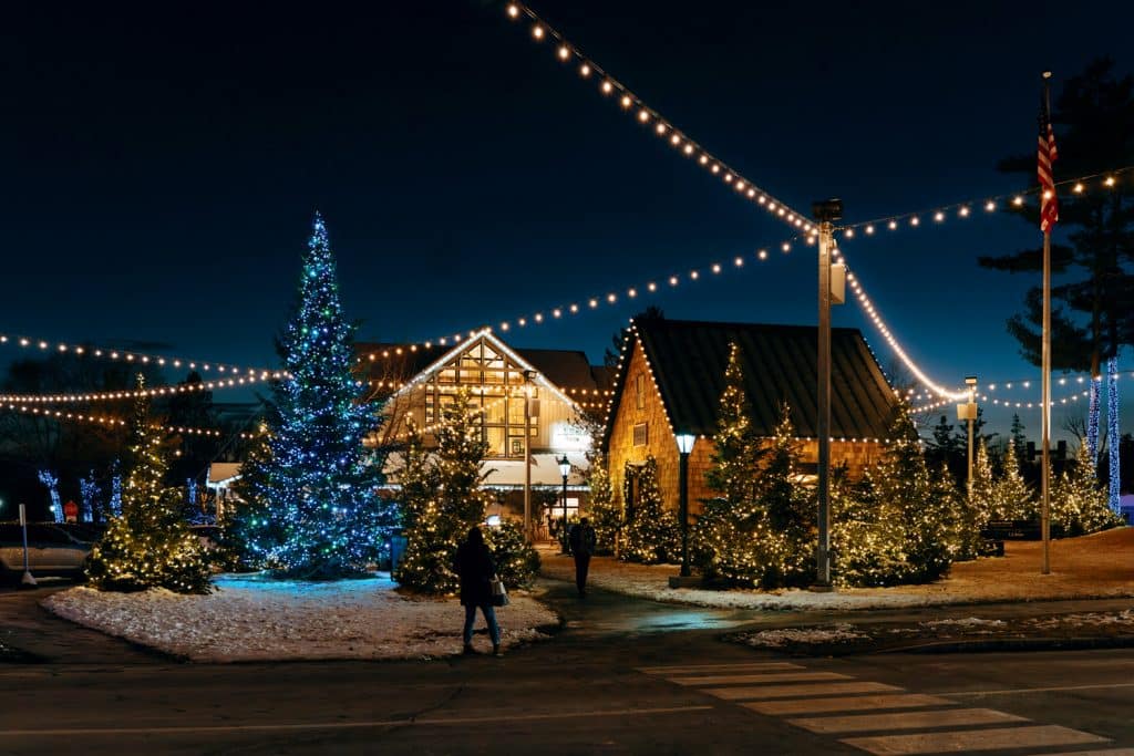 A Christmas tree adorned with blue lights stands amidst buildings decorated with white lights. People walk on a path under string lights in a snowy setting at night.