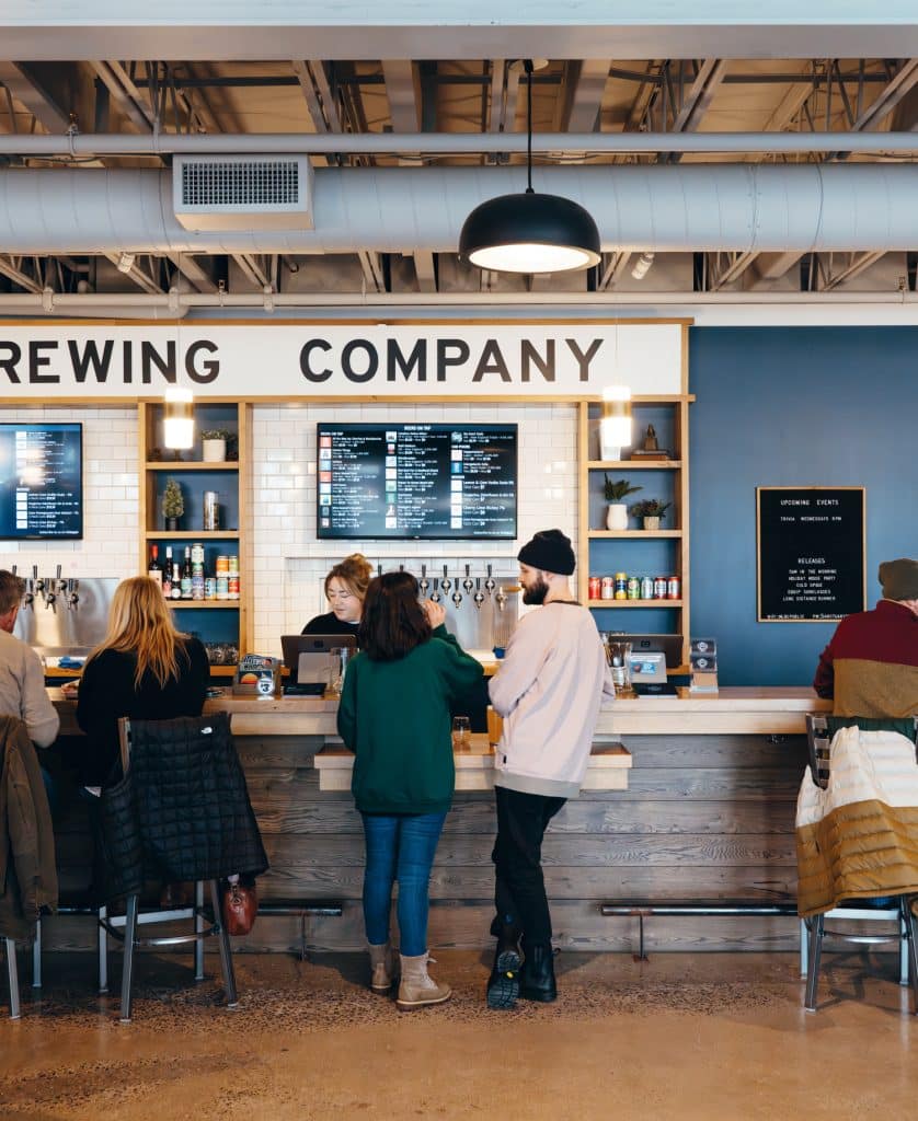 Customers standing at a brewery bar counter, with a staff member serving drinks. Menu boards are displayed on the wall behind them.