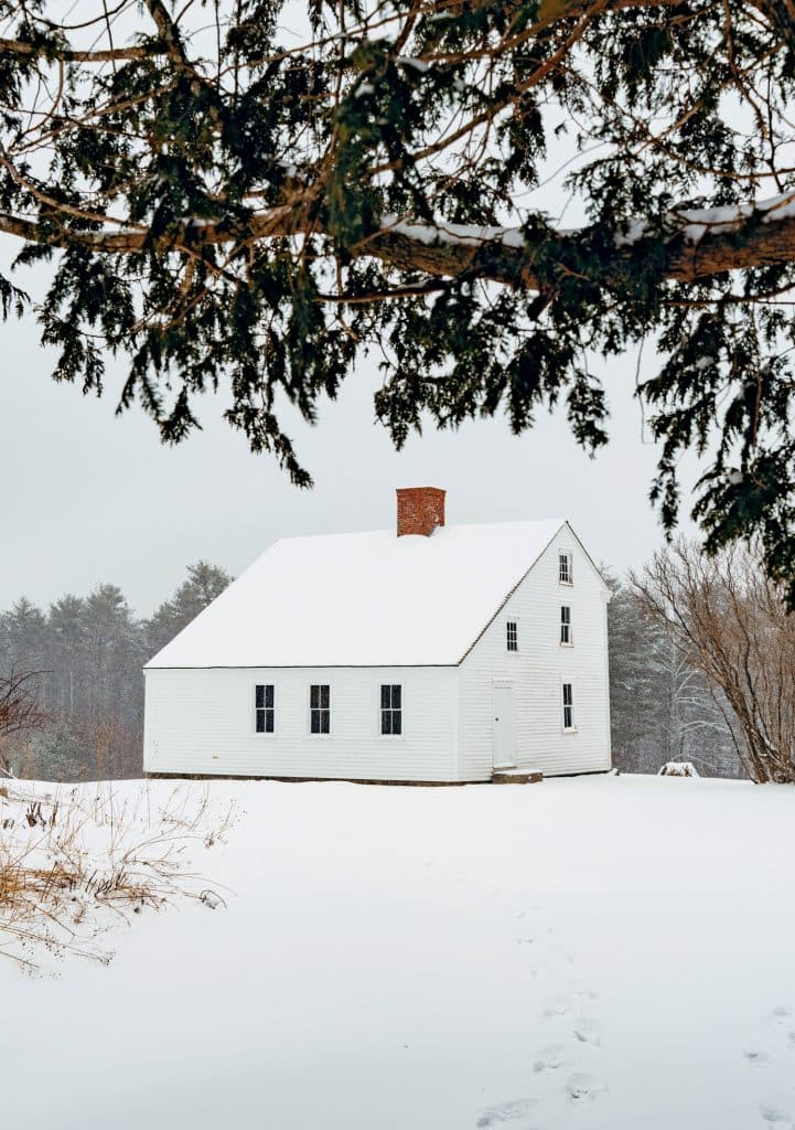 A white house with a chimney stands in a snowy landscape, surrounded by leafless trees and a pine branch in the foreground.