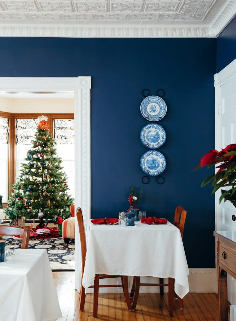 Dining room with blue walls, decorated with blue plates. Table set with red napkins. Christmas tree in the adjacent room.