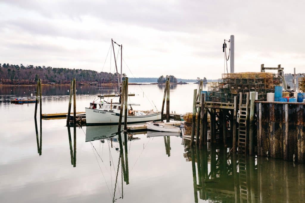 A fishing boat docked at a calm, reflective waterfront. Lobster traps and other fishing equipment are stacked on the pier under a cloudy sky.