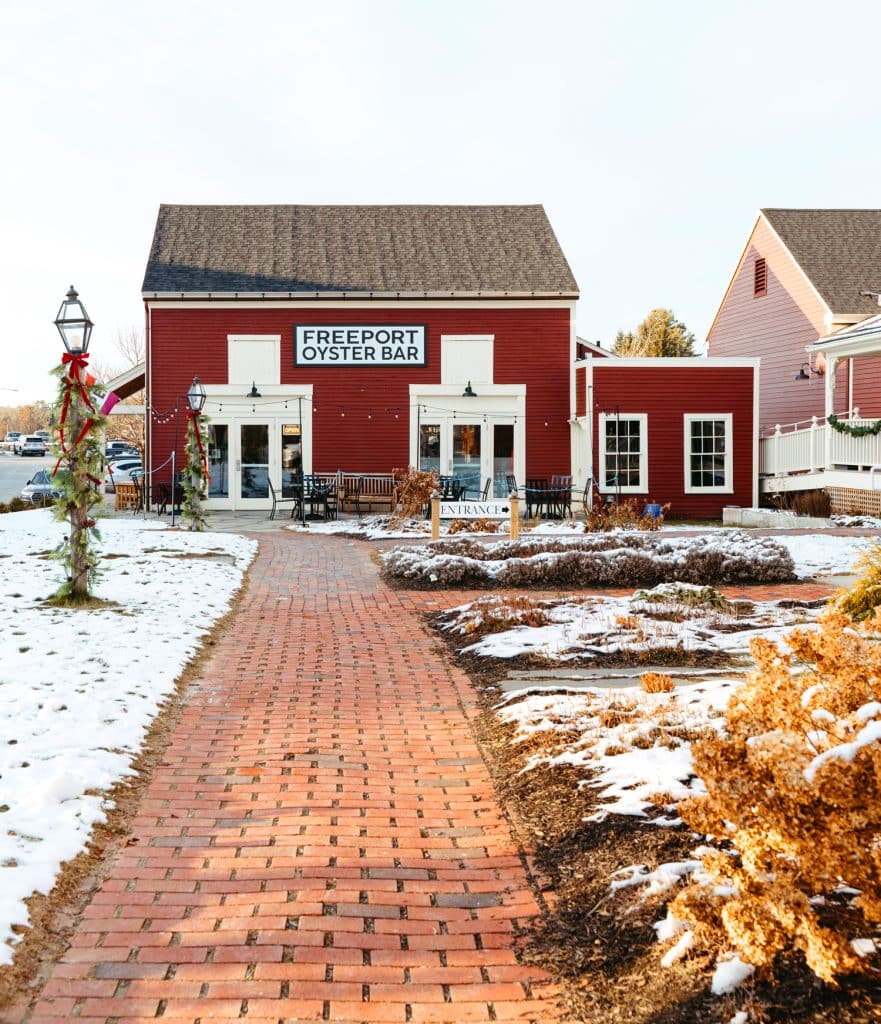 A red building with a sign reading "Freeport Oyster Bar." A brick walkway leads to the entrance, and snow is on the ground. Adjacent buildings and a lamppost are decorated for the holidays.