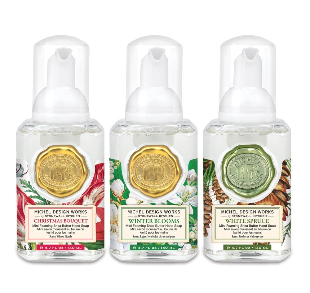 Three bottles of Michel Design Works foaming hand soap in Christmas-themed scents: Christmas Bouquet, Winter Blooms, and White Spruce, each with decorative labels and gold or green seals.