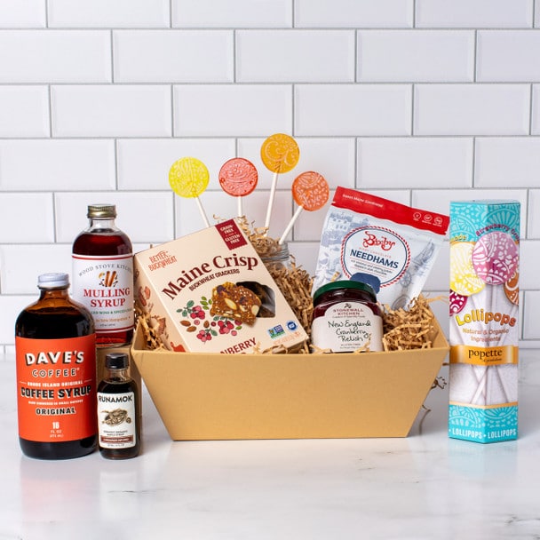 A gift basket containing coffee syrup, mulling syrup, berry crisps, needhams, chocolate wafer cookies, maple syrup, and colorful lollipops is displayed on a tiled background.