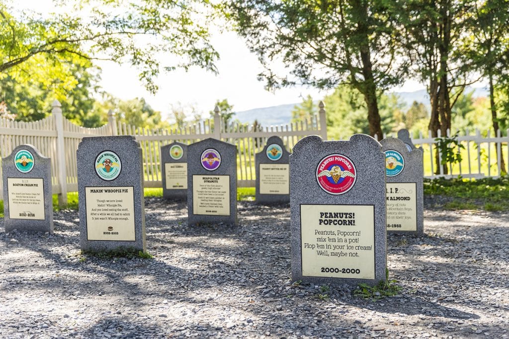 Several Ben & Jerry's flavor grave markers in a sunny, fenced yard, each with colorful logos and inscriptions.