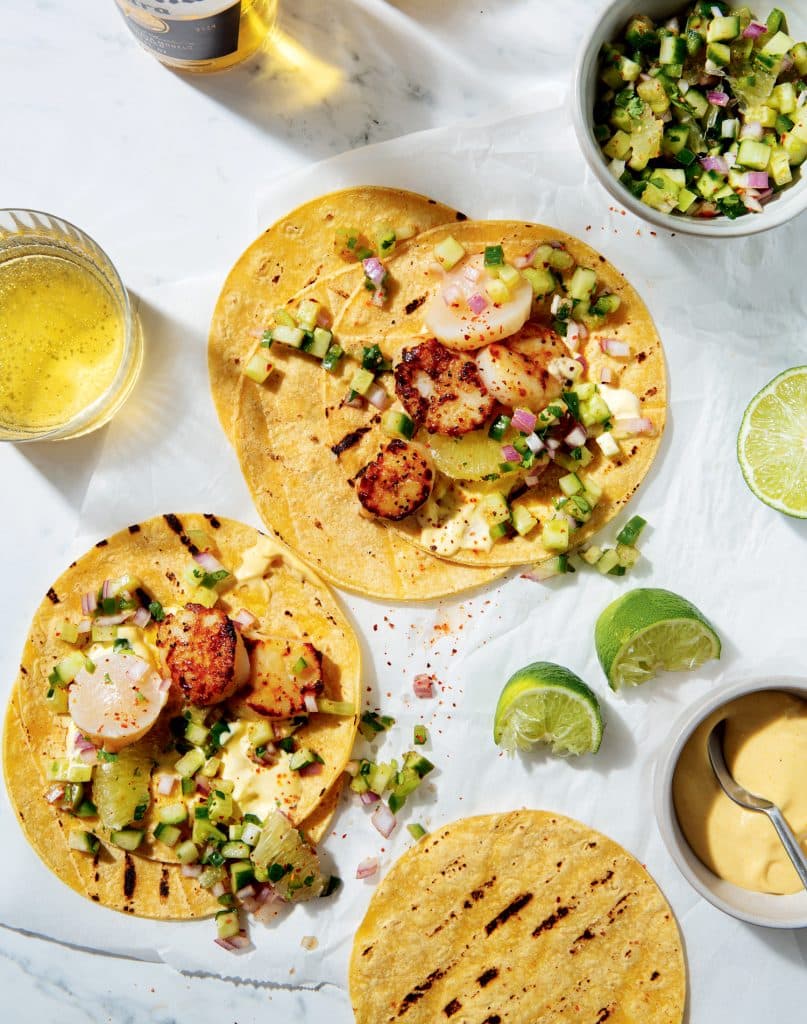 Grilled tortillas topped with seared scallops, diced vegetables, and lime halves on the side, accompanied by drinks.