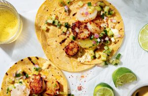 Two corn tortillas topped with grilled scallops, diced cucumber, onion, and salsa, alongside lime wedges and a glass of light beer.