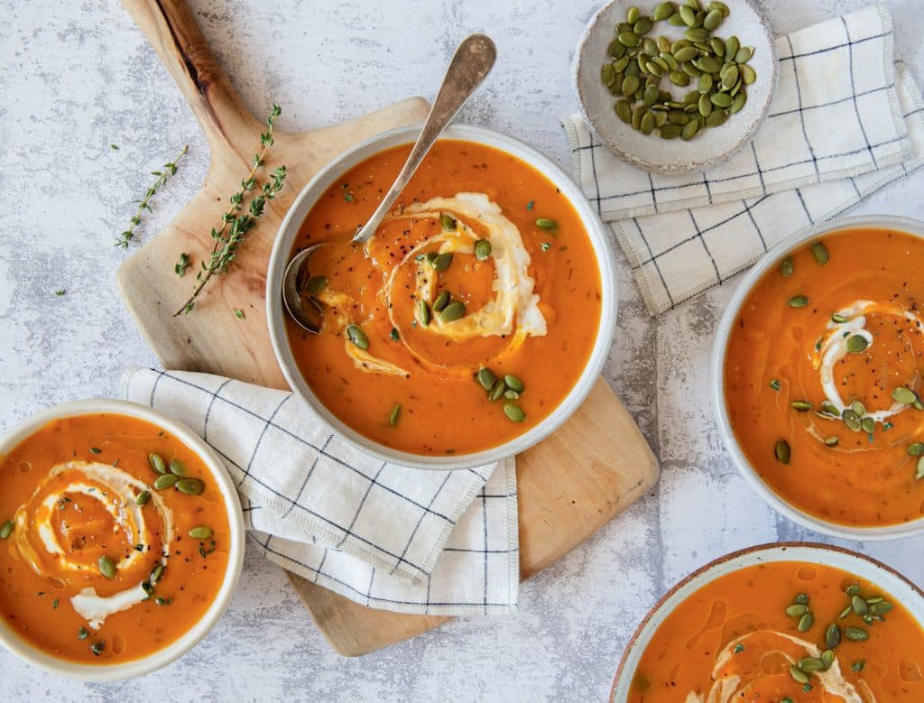 Bowls of pumpkin soup garnished with cream and pumpkin seeds on a table with a wooden board and cloth napkins.