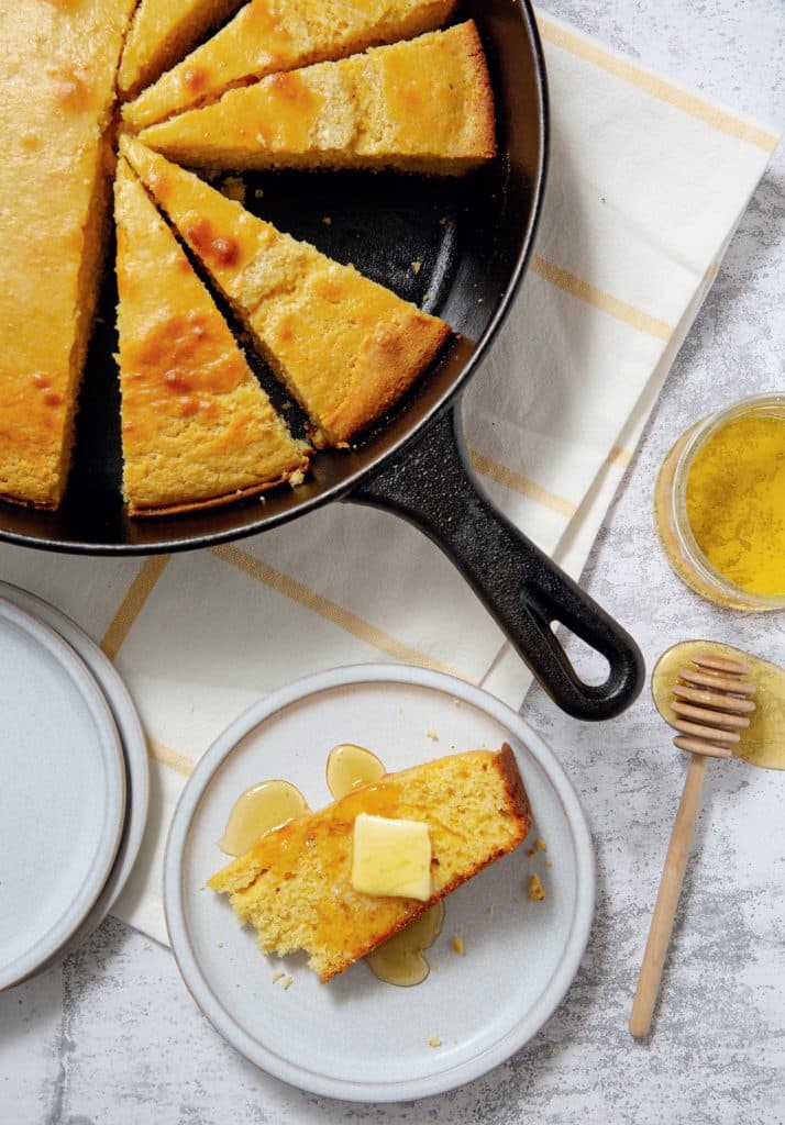 Skillet cornbread cut into wedges, with one piece on a plate topped with butter and honey. Nearby are a honey dipper, jar of honey, and two empty plates on a checkered cloth.