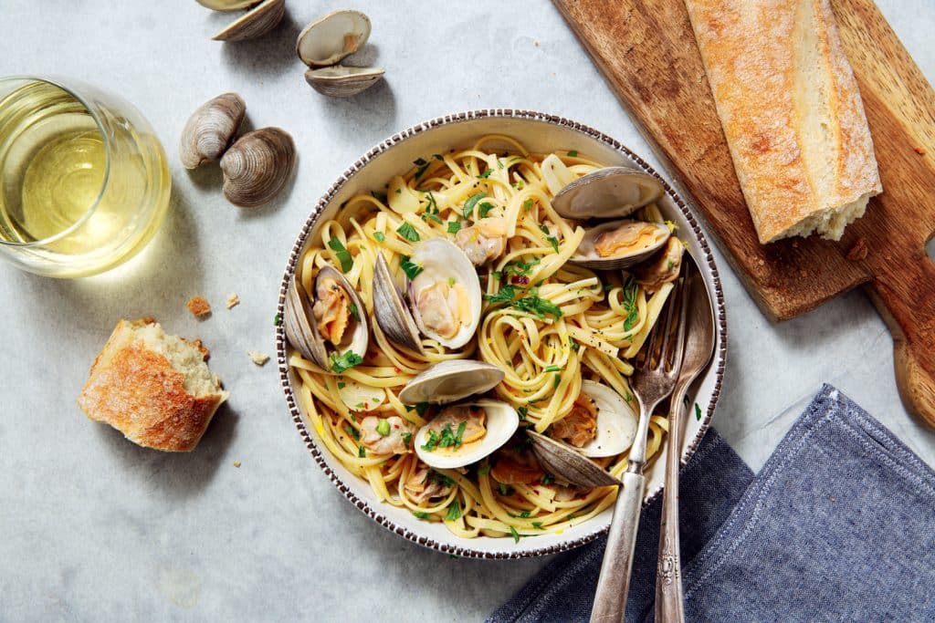 A bowl of clam linguine with parsley garnish, surrounded by a glass of white wine, clams, sliced baguette, and a fork and spoon on a gray surface with a napkin.