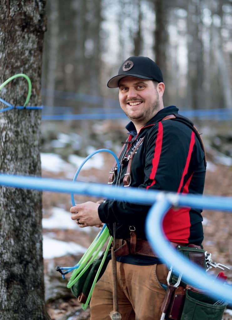 Person wearing a cap sets up blue tubing around trees in a wooded area with patches of snow.