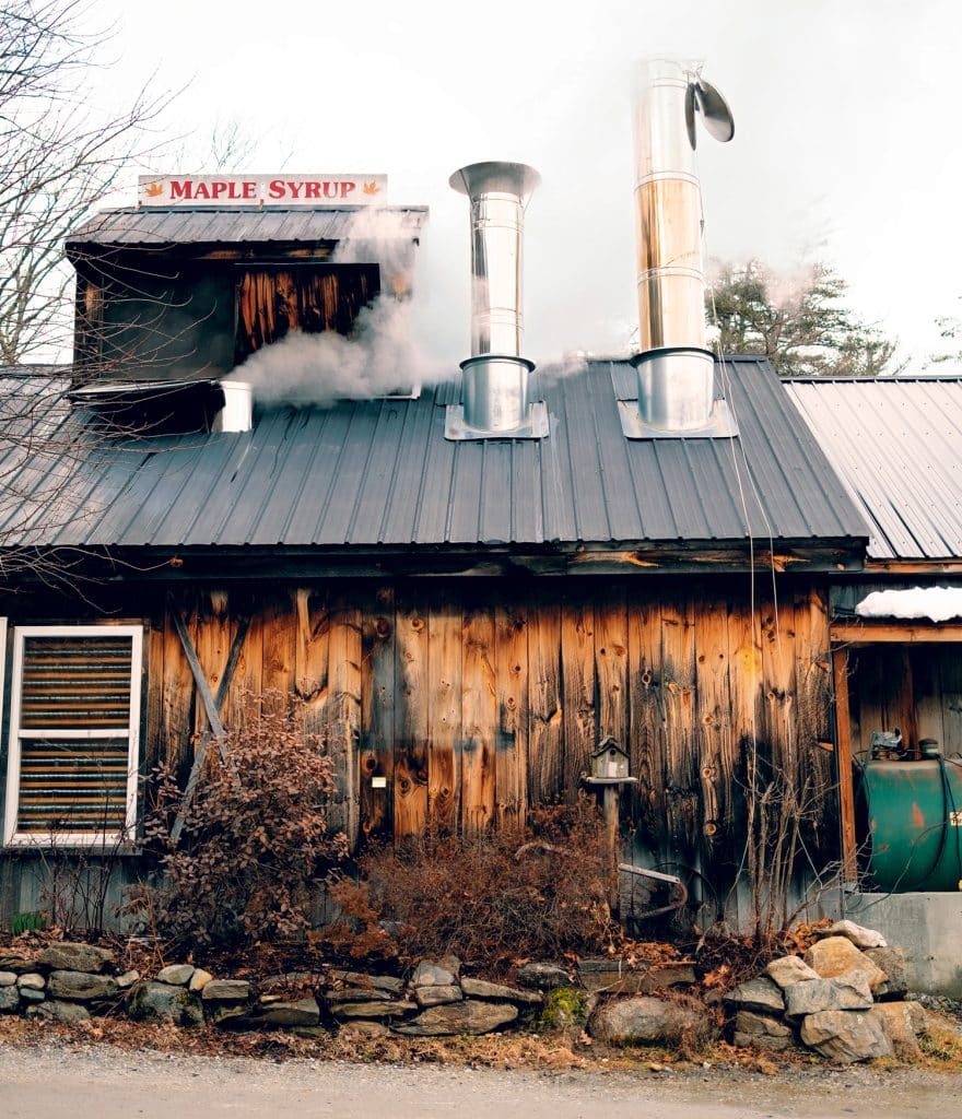 Rustic wooden building with "Maple Syrup" sign, two metal chimneys emitting steam, and surrounded by bare trees and stone accents.