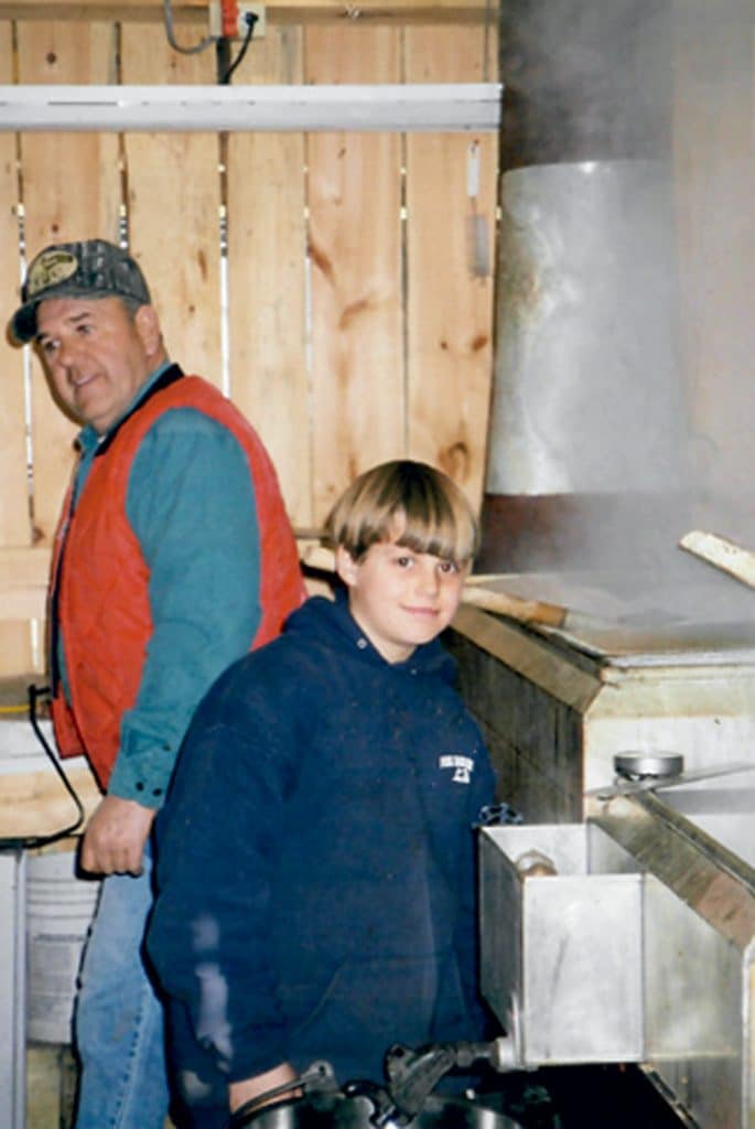 A man and a boy stand near a wooden wall in a room filled with steam, next to metal equipment. The man wears a cap and red vest; the boy is in a dark hoodie.