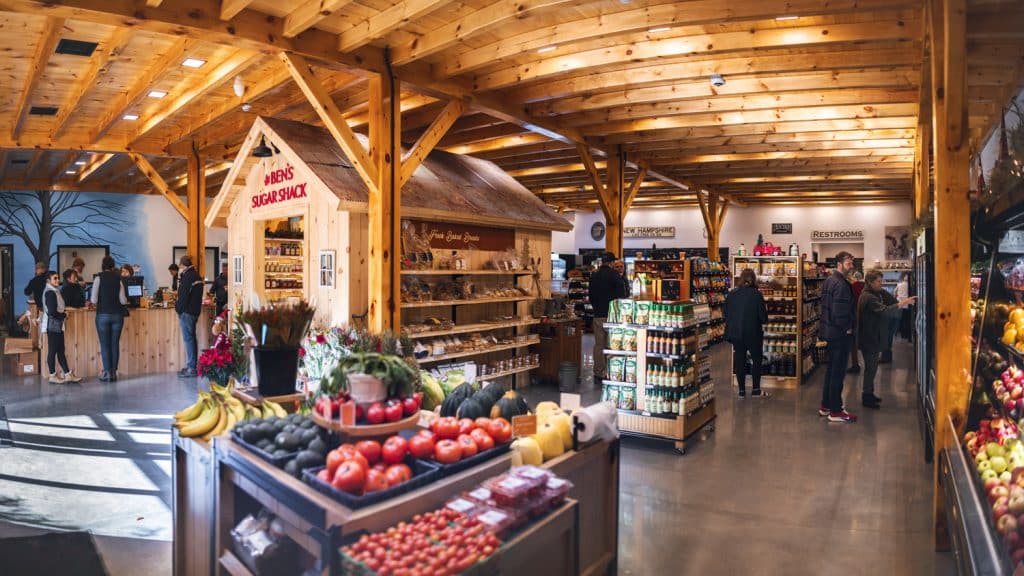 Interior of a wooden farm market with fresh produce, canned goods, and signs for restrooms. Shoppers browse various sections.