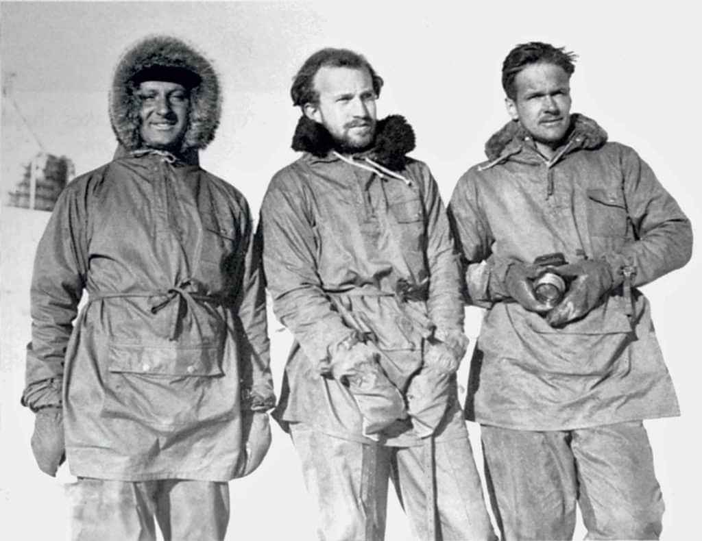 Three explorers in fur-lined coats stand together in snowy conditions, wearing gloves and warm clothing, with one holding a camera.