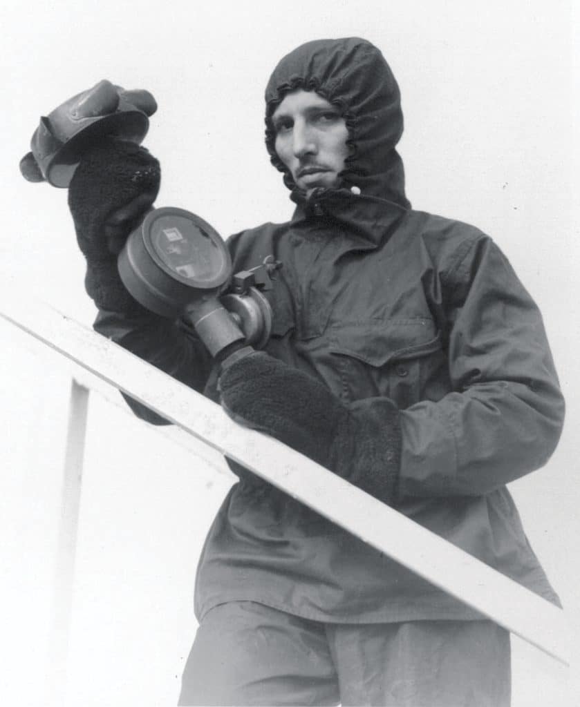 Person wearing a hooded jacket and gloves holds a gas mask, standing by a white railing.