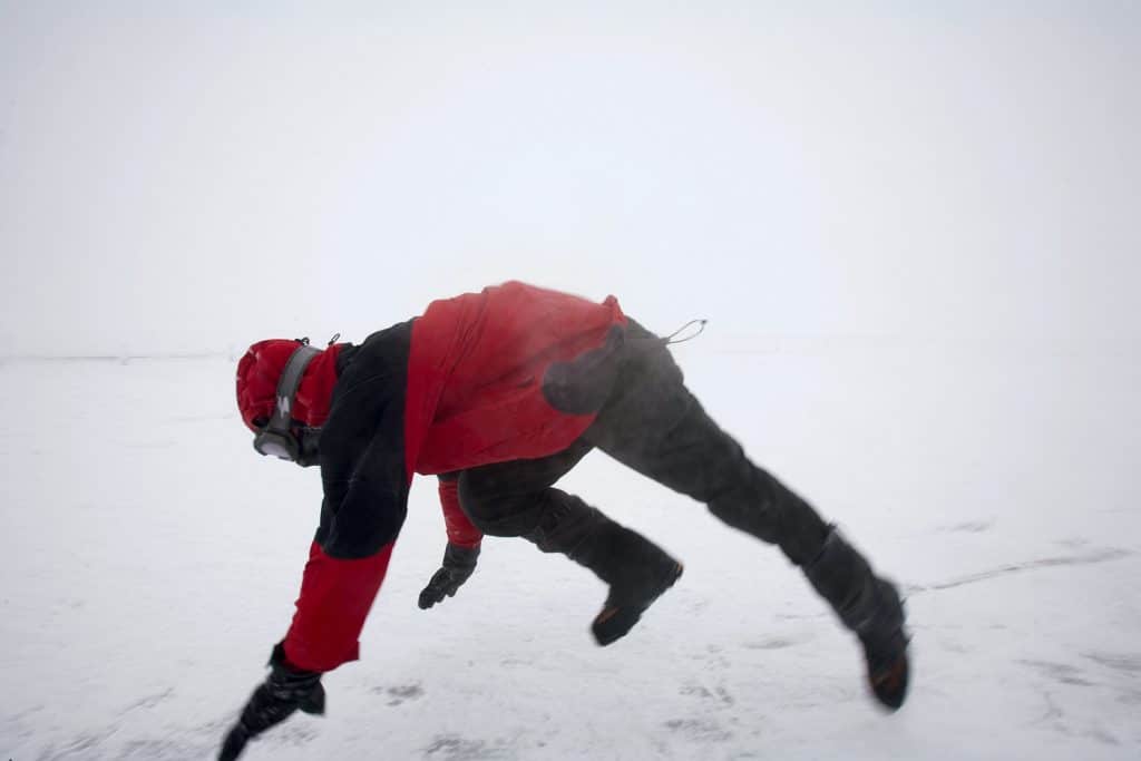 Person in red jacket leaning into strong winds on a snowy, foggy landscape.