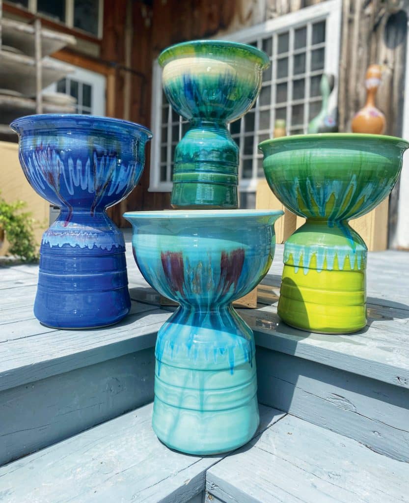 Four colorful ceramic vases with unique, round shapes and vibrant patterns are displayed on blue wooden steps outdoors.