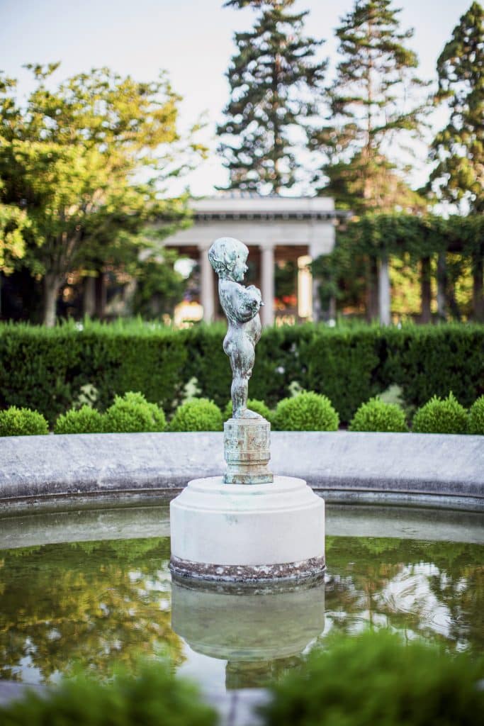 Statue of a child holding a jug in the center of a circular fountain, surrounded by greenery and trees, with a classical building in the background.