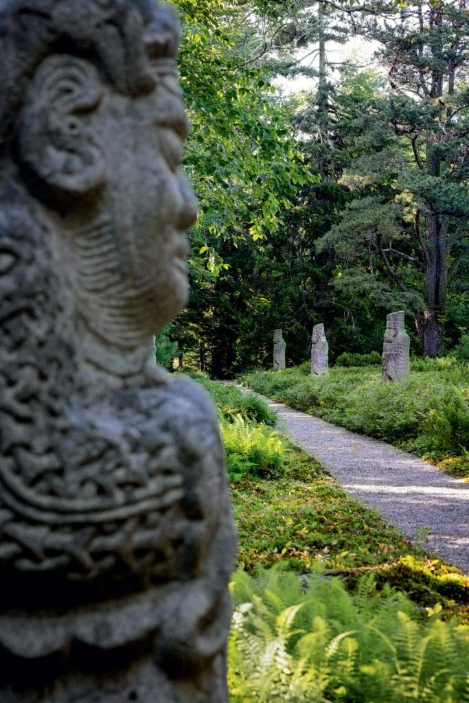 Stone statue in the foreground with a path leading through a lush, green forest scattered with more stone statues in the background.