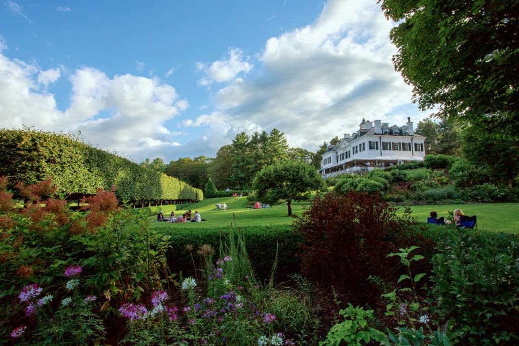 A large white house stands amidst a lush garden with blooming flowers and shrubs. People are sitting and relaxing on the lawn under a partly cloudy sky.