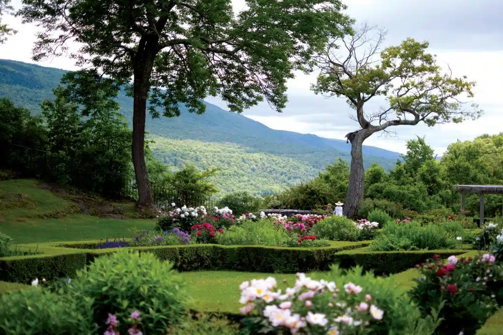 A scenic garden with colorful flowers and neatly trimmed hedges, framed by two large trees. In the background, rolling hills and a cloudy sky are visible.