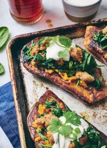 Close-up of baked sweet potatoes topped with spinach, crumbled sausage, sour cream, and cilantro on a baking sheet. Drinks and a cloth are visible nearby.