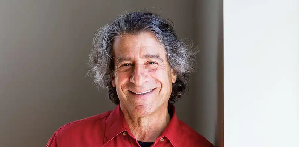 A smiling man with gray hair, wearing a red shirt, stands against a light-colored wall.