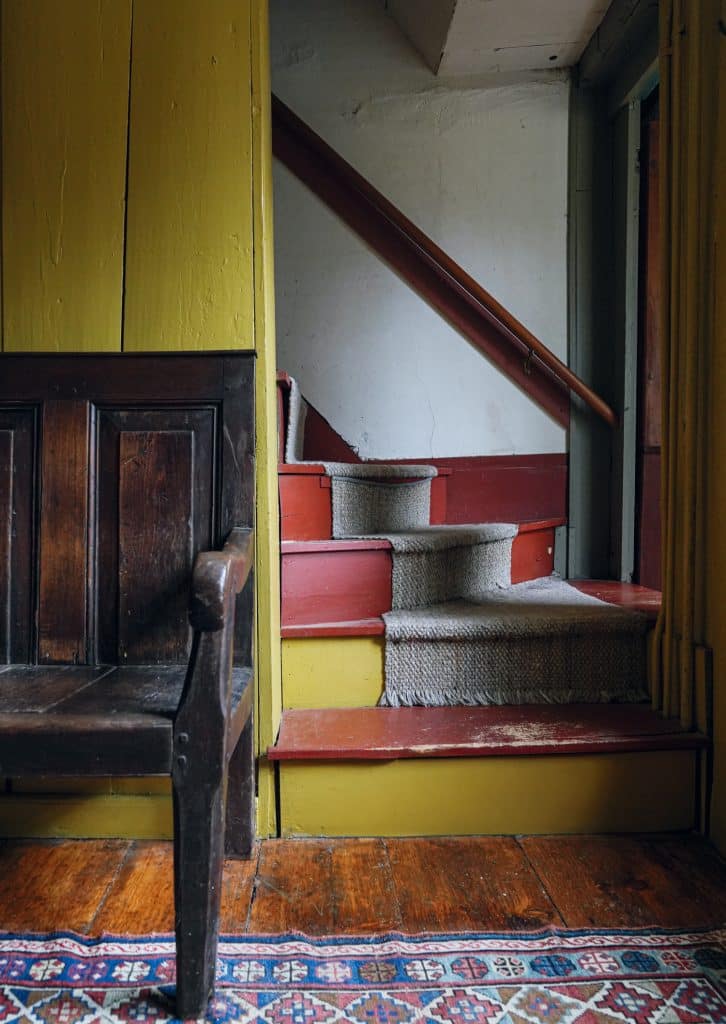 A wooden bench sits beside a staircase with red steps and a tan carpet runner, against a yellow wall. A patterned rug is on the wooden floor.