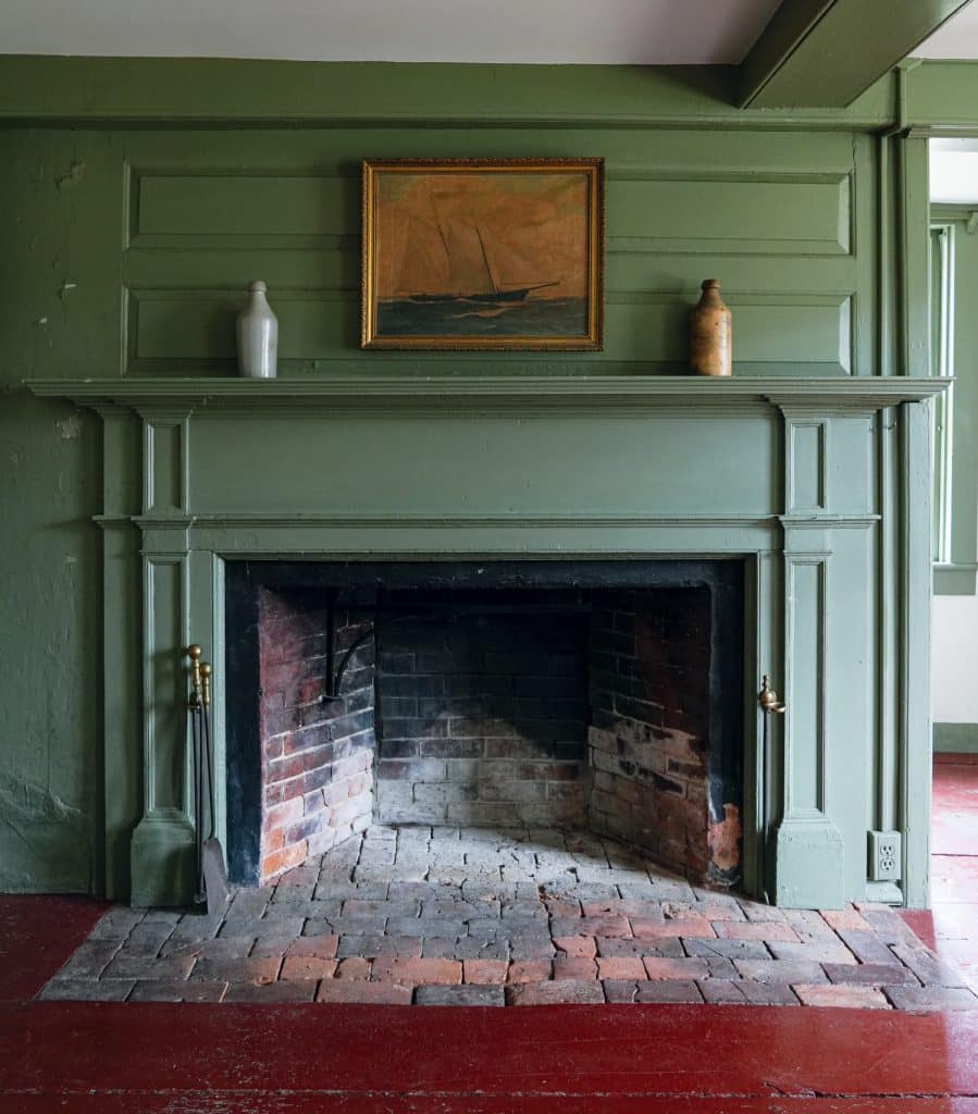 Green-painted fireplace with a brick hearth, flanked by two containers. A framed sailboat painting is centered above. Floor is red brick. Multiple layers of paint are visible on the left wall.